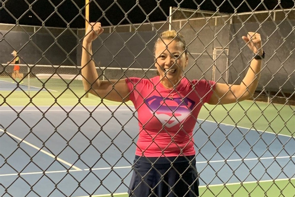 Woman flexing arms behind chain-link fence on tennis court.